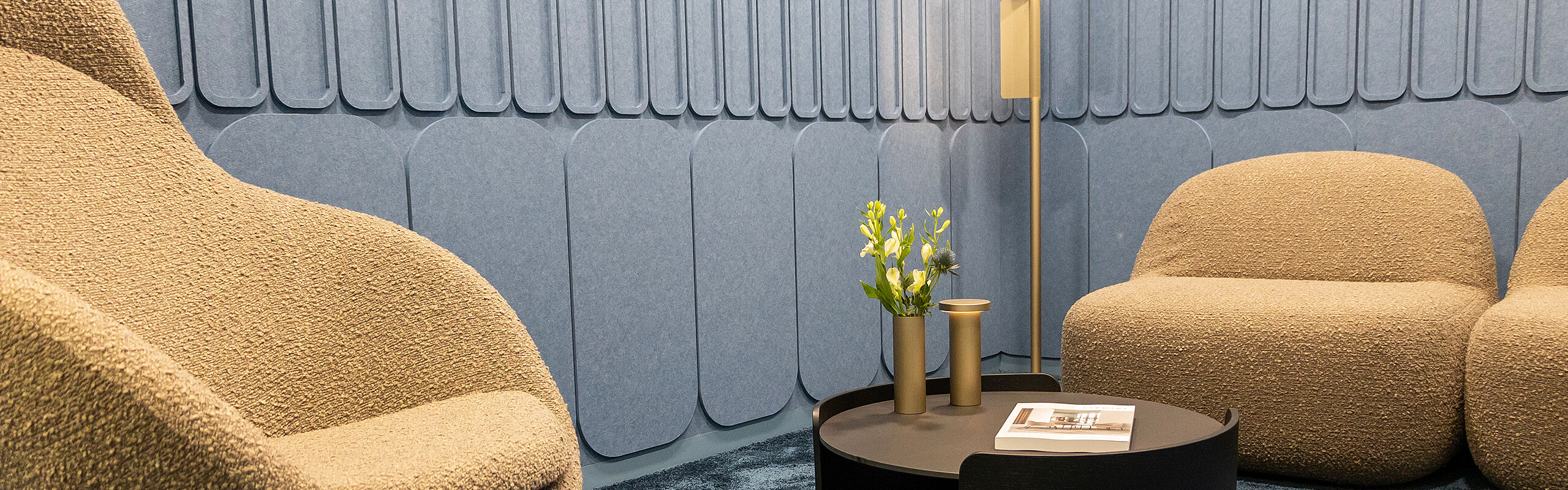 Cozy office lounge area with upholstered armchairs, a round table and decorative lighting in front of an acoustically designed wall.