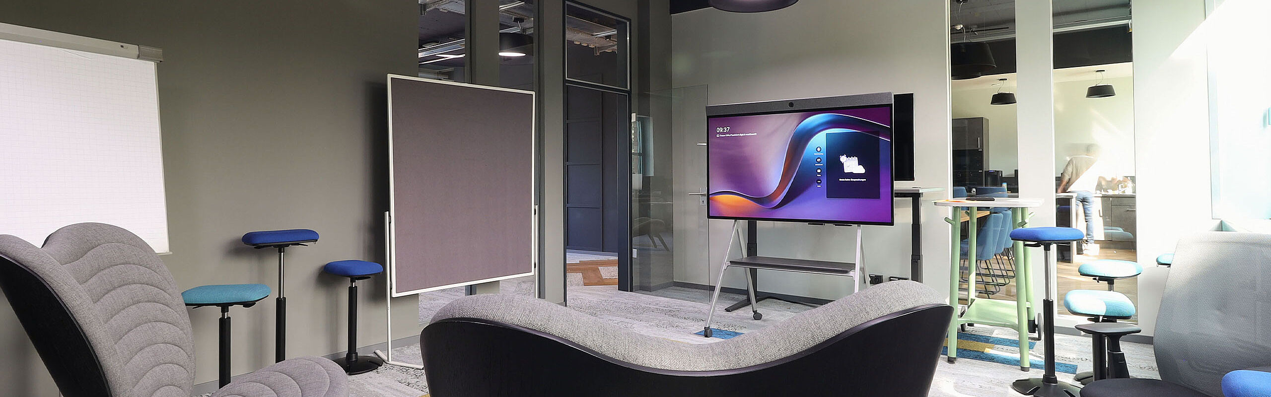 Kreativer Meetingraum mit Lounge-Sesseln und mobilen Hockern; in der Mitte ein großer Bildschirm auf Stativ, daneben Whiteboard und Stehtische vor Glaswänden.