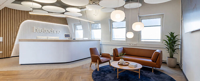  A bright reception area by Bio-Techne featuring curved wood paneling, a seating area, and pendant lights.
