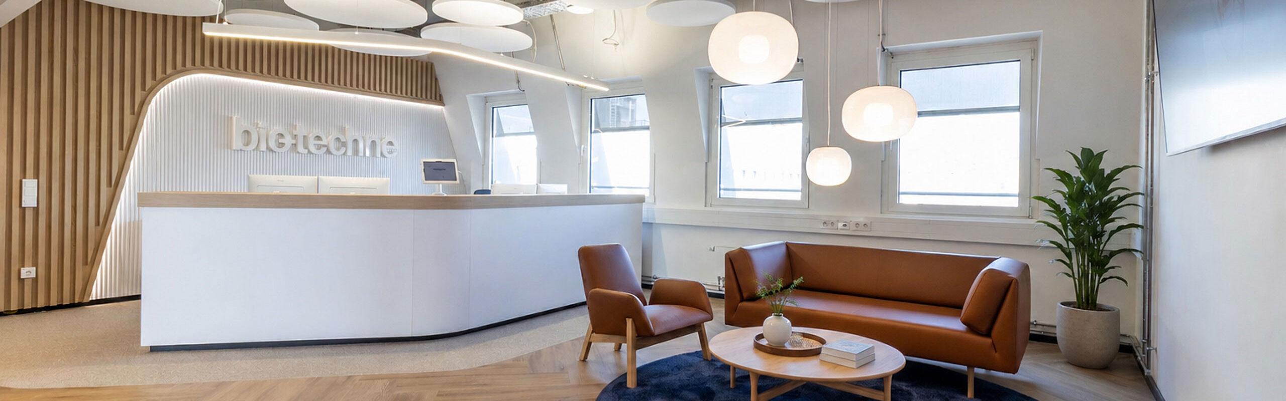  A bright reception area by Bio-Techne featuring curved wood paneling, a seating area, and pendant lights.