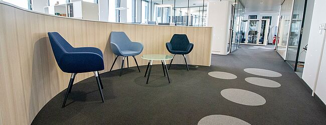 Eine Ecke mit einem kleinen Tisch und drei modernen blauen Stühlen, die an einer halbrunden Wand stehen und zu der ein Durchgang führt.