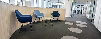Eine Ecke mit einem kleinen Tisch und drei modernen blauen Stühlen, die an einer halbrunden Wand stehen und zu der ein Durchgang führt.