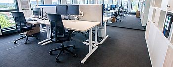Ein Bürotisch mit Monitoren, zwei schwarze Bürostühle und die übrigen Arbeitsbereiche im Hintergrund.
