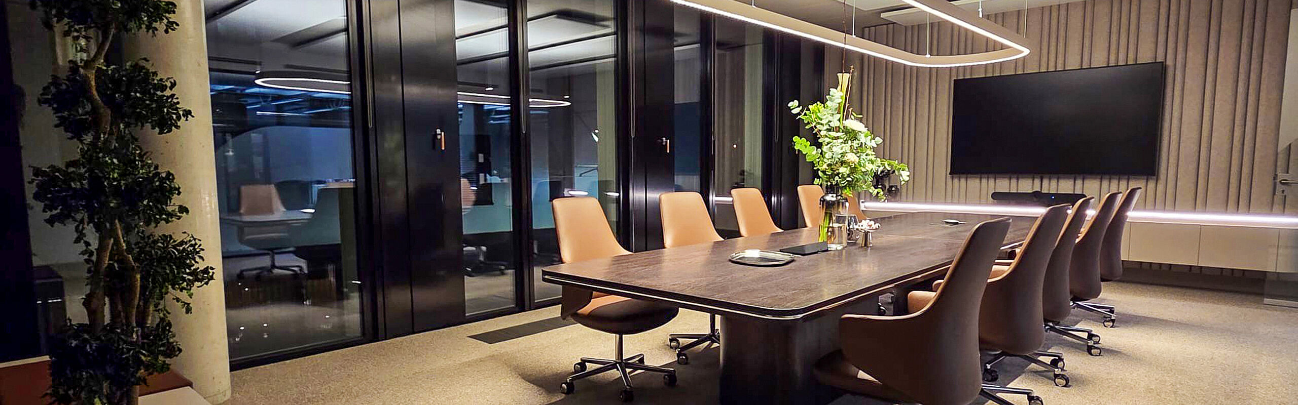 Hochwertiger Konferenzraum mit großem Holztisch, Lederstühlen, Bildschirm und markanter Designbeleuchtung in einer modernen Büroarchitektur.