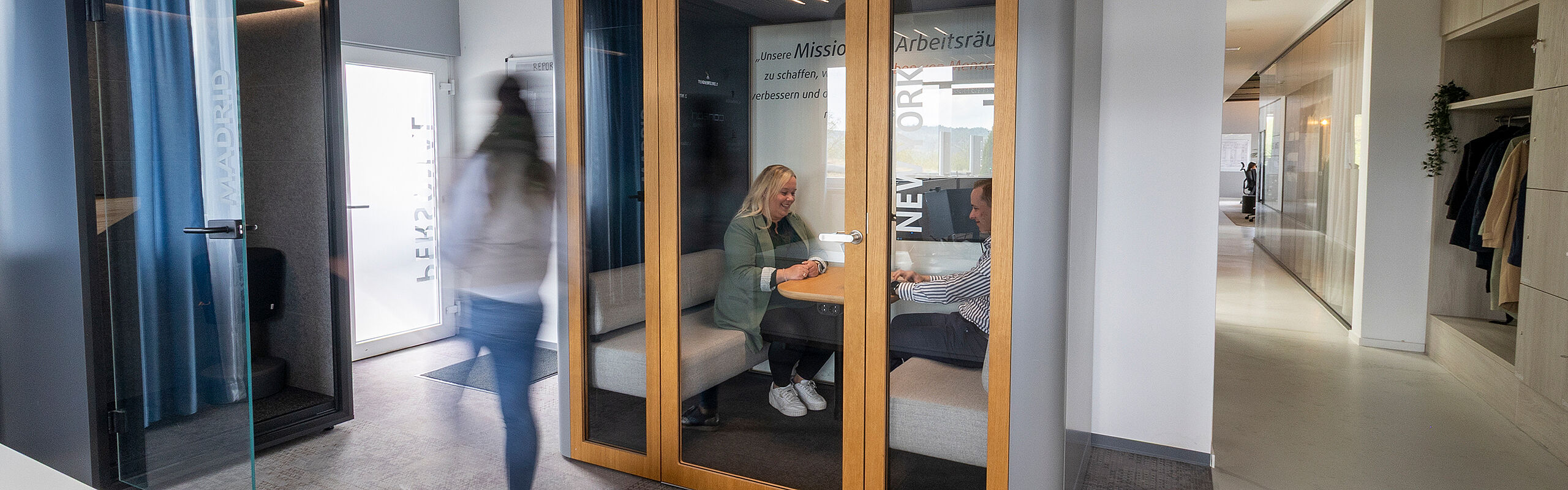 Eine Person läuft auf eine Telefonkabine zu zwei andere Personen mittig im Bild sitzen in einer Akustikbox rechts im Bild sieht man den Flur eines Büros mit Garderobe