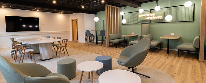 Modern breakout and collaboration area in a contemporary office setting, featuring a central meeting table, lounge seating, high tables, and semi-private booth areas, designed to support informal exchange, teamwork, and flexible use.