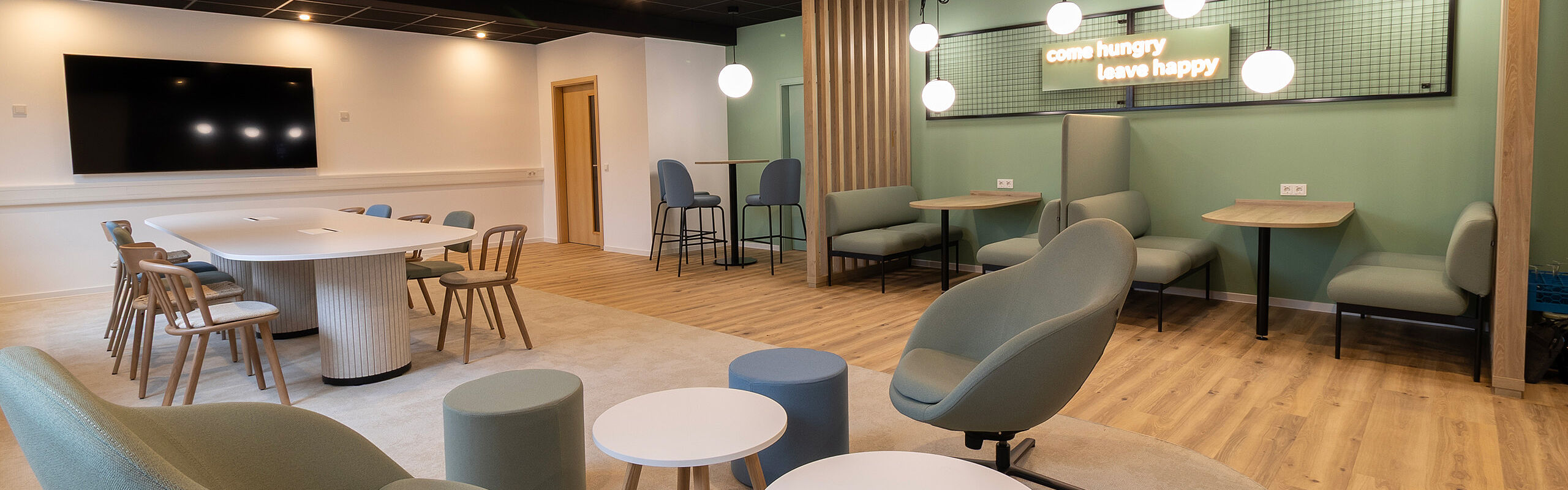 Modern breakout and collaboration area in a contemporary office setting, featuring a central meeting table, lounge seating, high tables, and semi-private booth areas, designed to support informal exchange, teamwork, and flexible use.