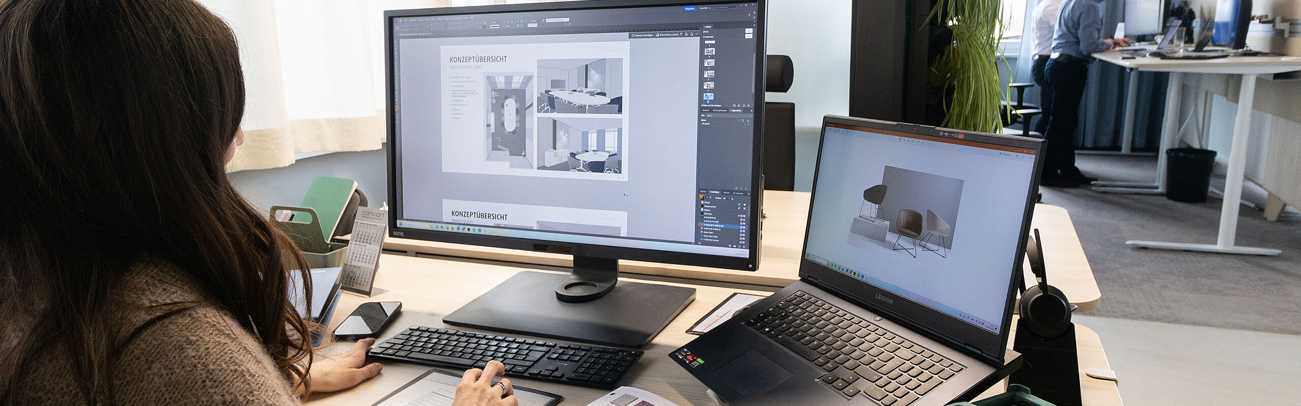 Mitarbeiterin arbeitet am Schreibtisch mit großem Monitor und Laptop an einem Innenraumkonzept; auf dem Tisch liegen Material- und Farbmuster, im Hintergrund ein offener Bürobereich.