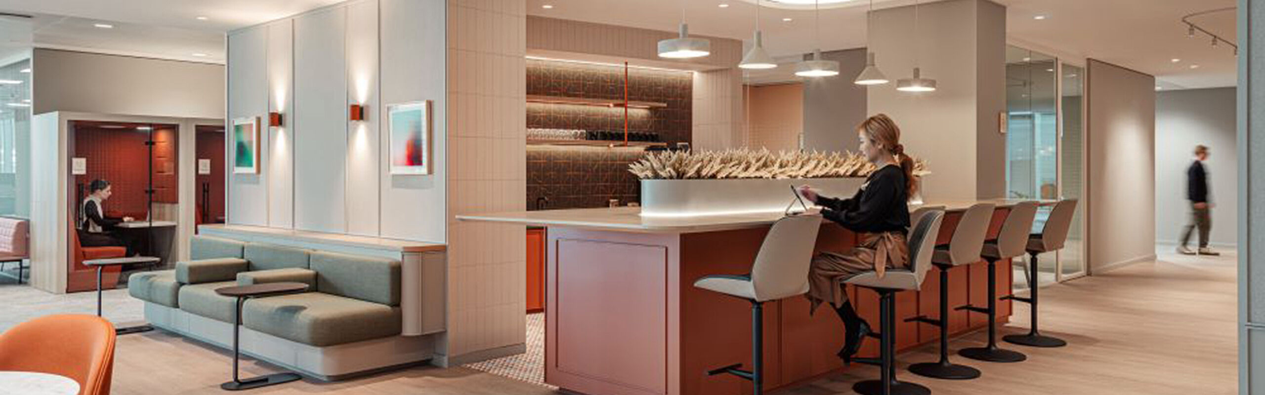 Moderne Büro-Lounge mit Barbereich und Sitzgelegenheiten, eine Person arbeitet am Laptop an einem Hochtisch in einer offenen, warm gestalteten Arbeitsumgebung.