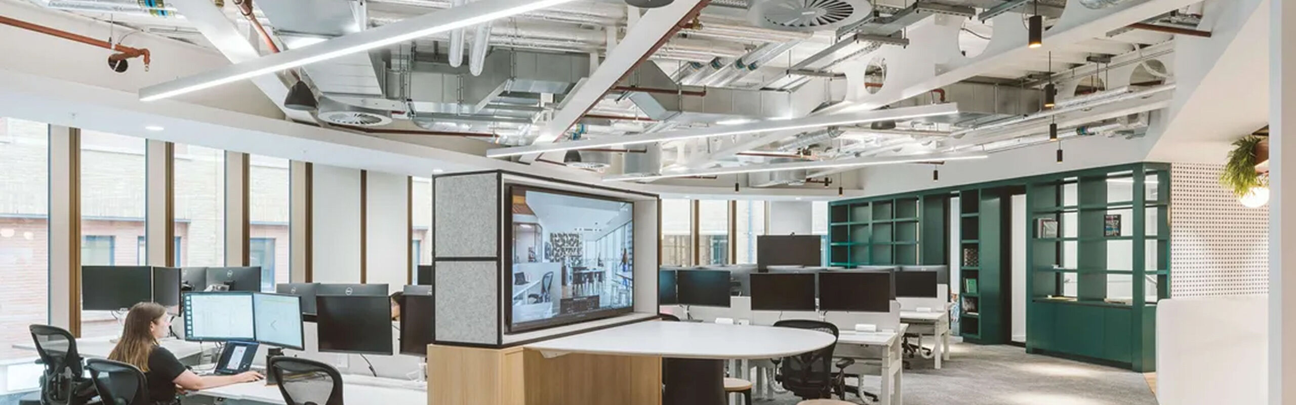 Modernes, offenes Büro mit mehreren Bildschirmarbeitsplätzen, zentralem Besprechungsbereich und grüner Raumstruktur, durchflutet von Tageslicht.