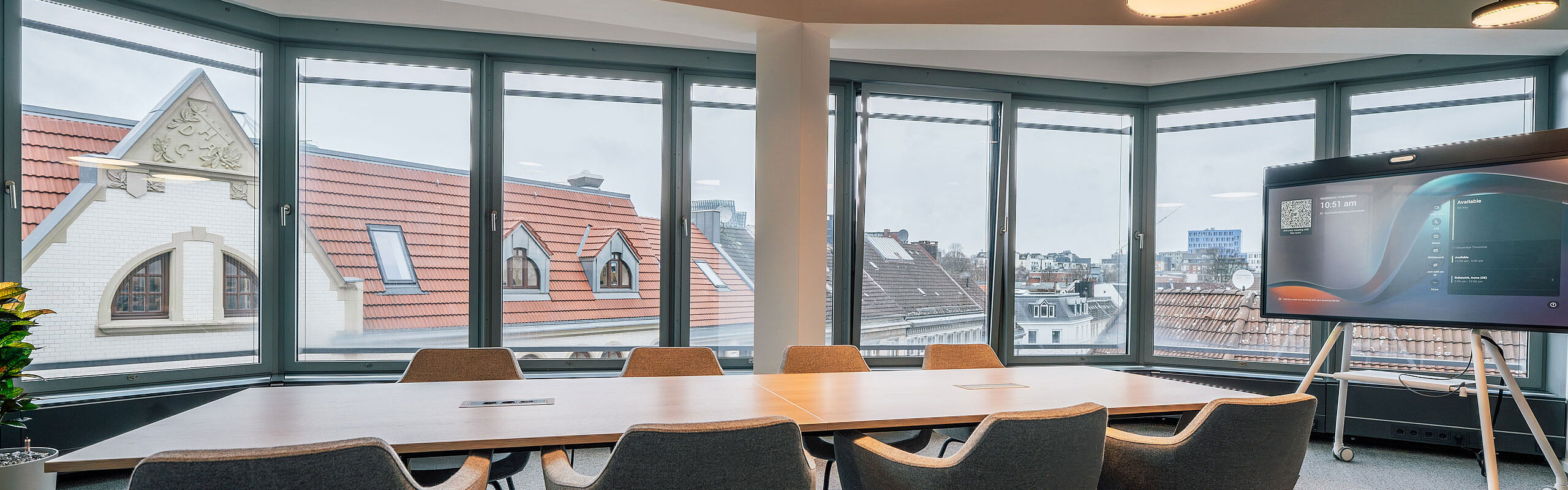 Moderner Konferenzraum mit großem Besprechungstisch, bequemen Sitzmöbeln und bodentiefen Fensterfronten mit Blick über die Dächer der Stadt.