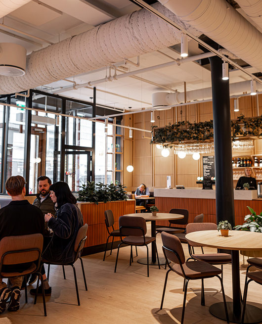 Offene Arbeits- und Aufenthaltsfläche mit Cafécharakter, sichtbarer Gebäudetechnik und vielfältigen Sitzbereichen – moderne Bürogestaltung für Austausch und informelle Zusammenarbeit.