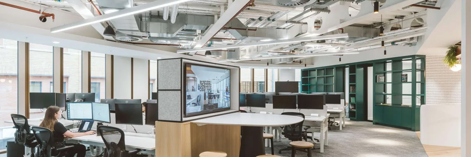 Modernes Open-Space-Büro mit Arbeitsplätzen, Monitoren und integrierter Medientechnik, offener Decke mit sichtbarer Haustechnik sowie Besprechungsbereich – Beispiel für einen zeitgemäßen Büroinnenausbau.