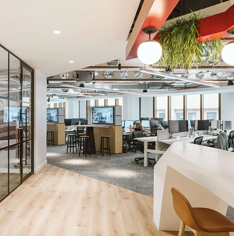 Modern gestaltetes Open-Space-Büro mit Arbeitsplätzen, Besprechungsinseln und integrierter Medientechnik, kombiniert mit Holz- und Teppichböden, offener Decke und begrünten Gestaltungselementen.
