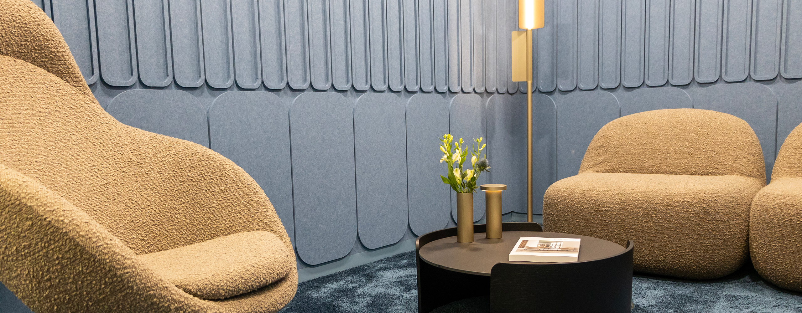 Gemütlicher Loungebereich im Büro mit gepolsterten Sesseln, rundem Tisch und dekorativer Beleuchtung vor einer akustisch gestalteten Wand.