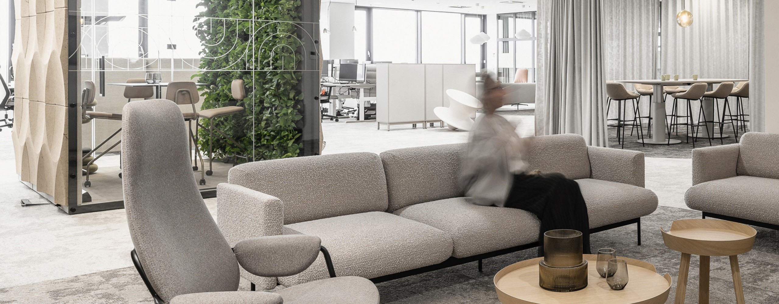 Offener Büro-Loungebereich mit Sofa, Sesseln und Couchtischen, kombiniert mit Arbeits- und Besprechungszonen sowie integrierter Begrünung.