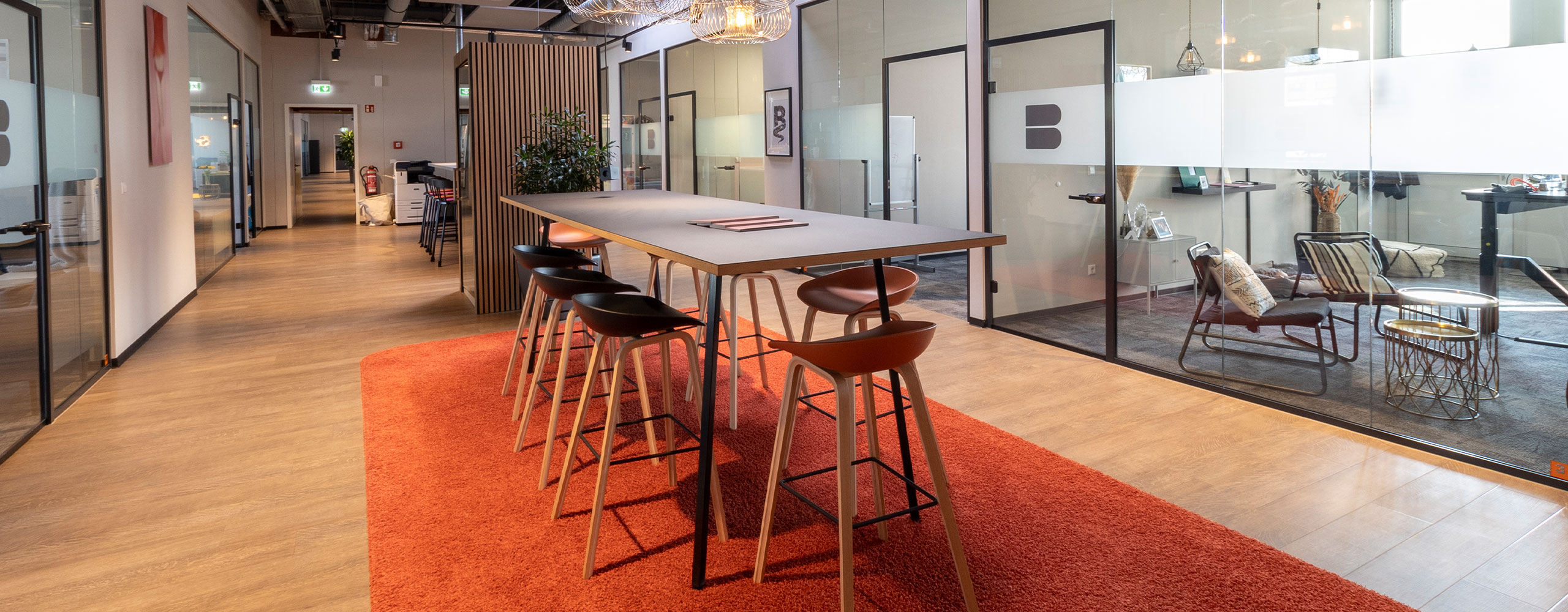 Offener Bürobereich mit hohem Besprechungstisch und Barhockern auf einem roten Teppich, umgeben von Glasbüros und Loungebereichen in einer modern gestalteten Arbeitsumgebung