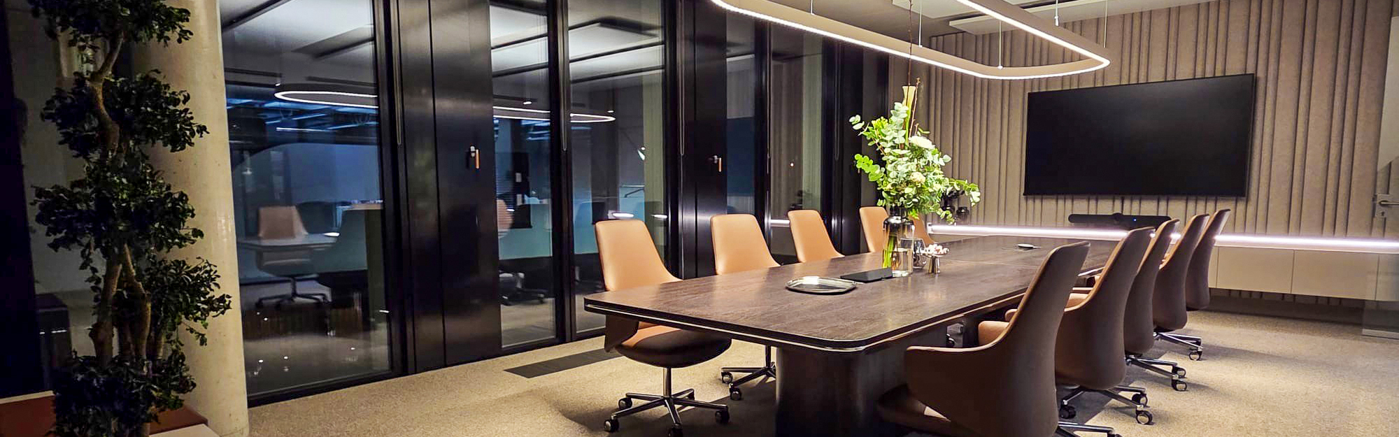 Modern conference room with a large meeting table, leather chairs, screen and design lighting in front of floor-to-ceiling glass windows.