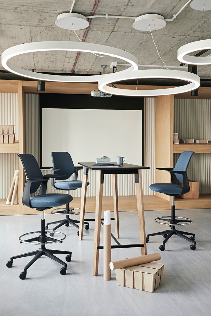 Modern workshop or meeting area with a high table, swivel chairs, presentation surface and distinctive ring lighting in an open workspace environment.