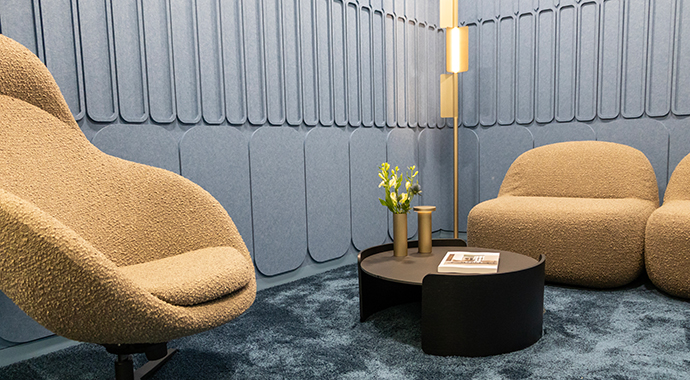 Gemütlicher Loungebereich im Büro mit zwei gepolsterten Sesseln, rundem Tisch und dekorativer Beleuchtung vor einer akustisch gestalteten Wand.