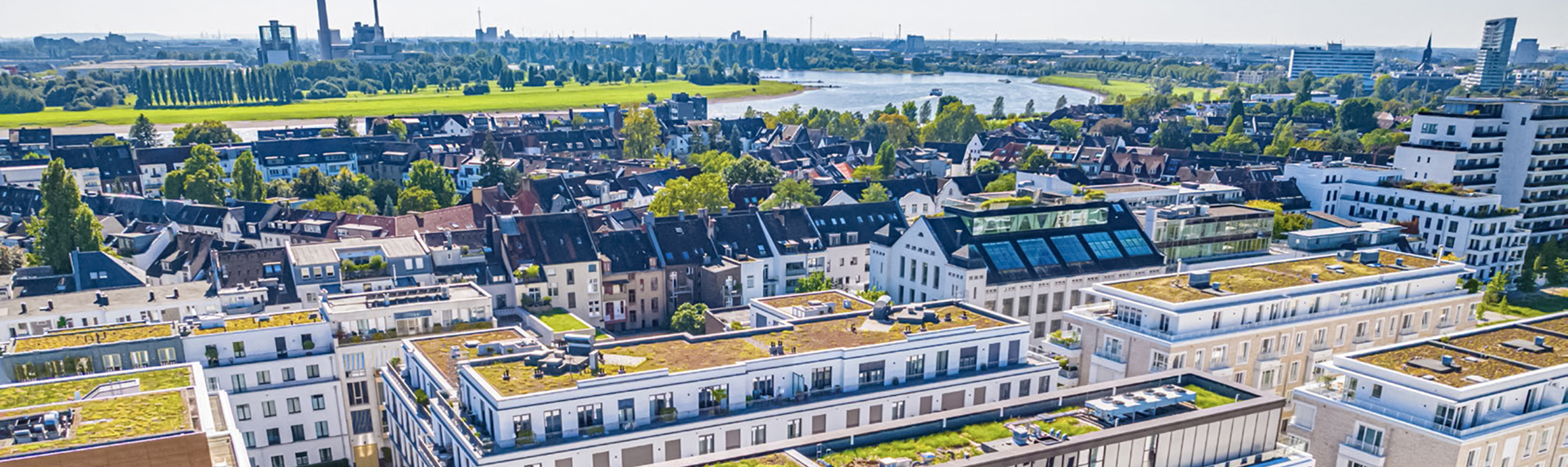Luftaufnahme eines urbanen Stadtviertels mit Wohngebäuden, begrünten Dächern und Flusslandschaft im Hintergrund.