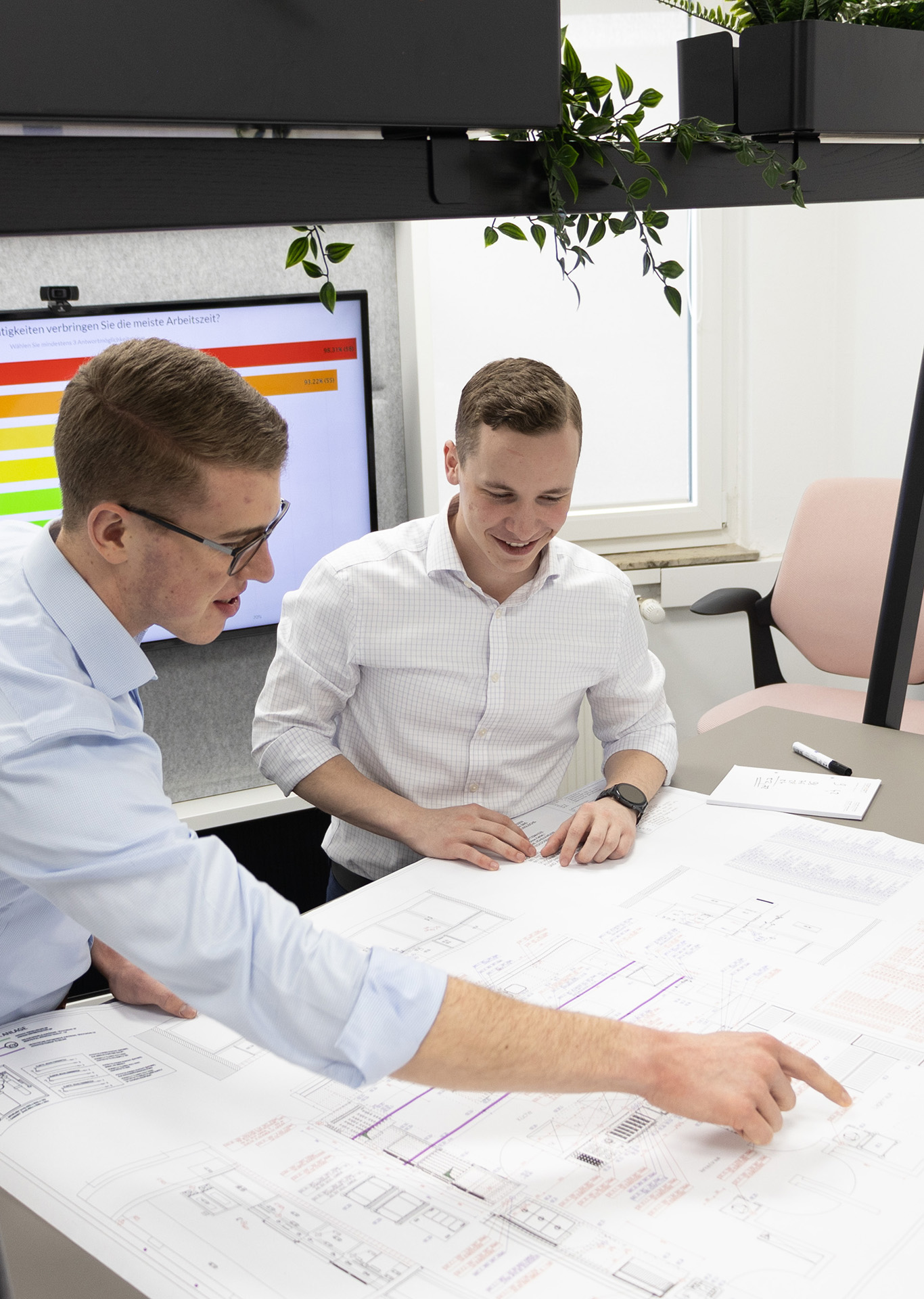 Zwei Personen schauen sich einen Gebäudeplan an im Hintergrund ist ein Bildschirm mit Diagramm