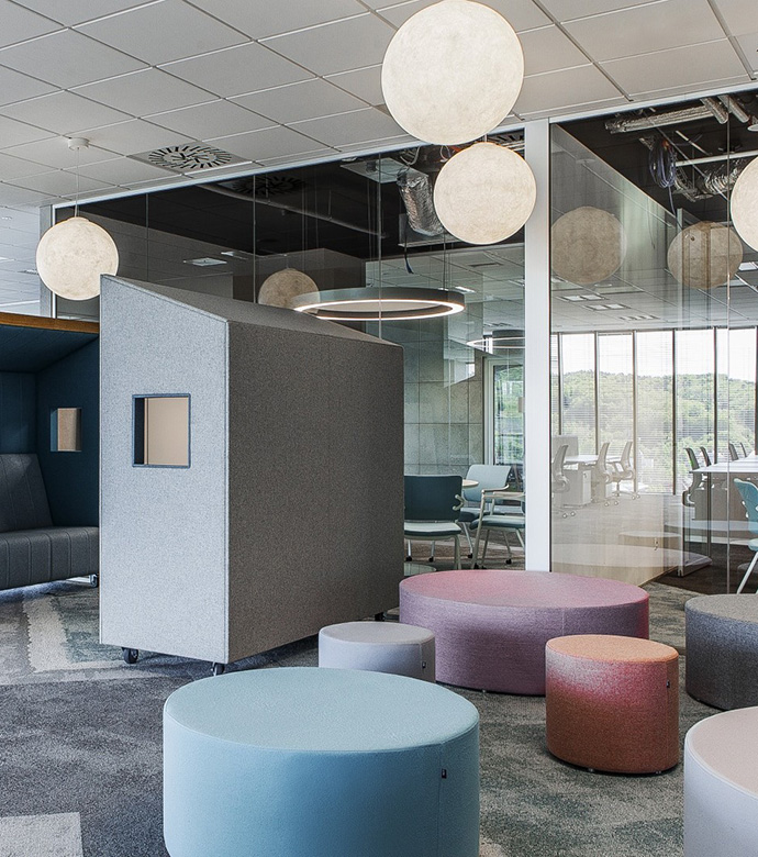 Modern office space with an acoustic seating booth, colorful poufs and glass meeting rooms, designed for flexible collaboration and retreat.