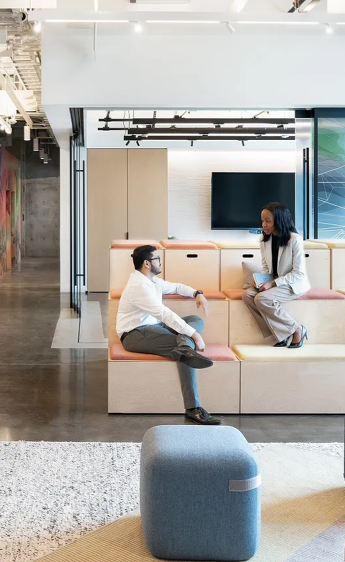 Two people in conversation in a modern office lounge with tiered seating, a screen and an open workspace environment.