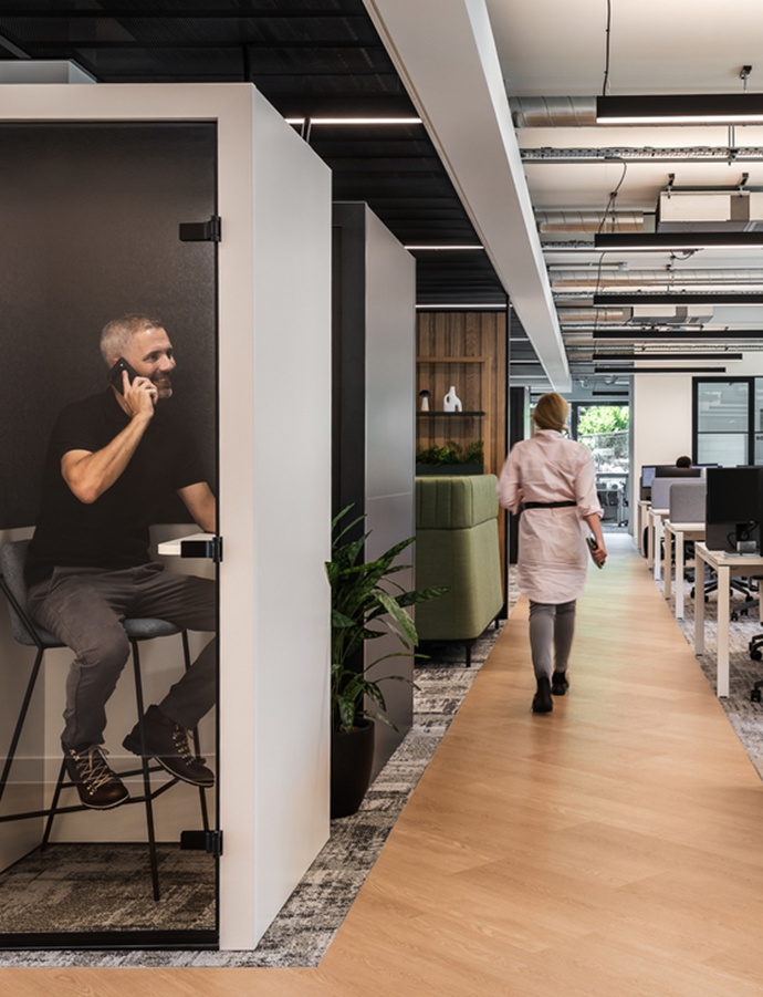 Modern office corridor with a phone booth for focused calls, open workstations and a person walking through a functional workplace environment.