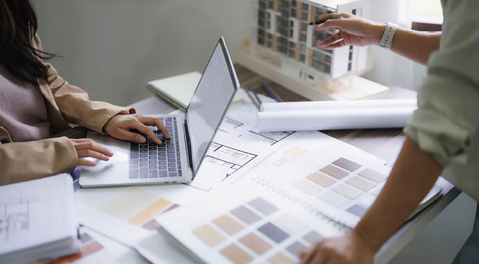 Auf einem Tisch liegen ein Laptop ein Farbprofile zwei Personen arbeiten daran