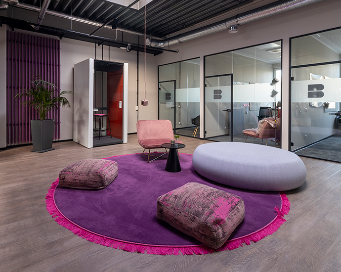 Bunte Sitzecke im Büro mit rundem lilanen Teppich Sitzkissen und Glaswänden mit Büros im Hintergrund