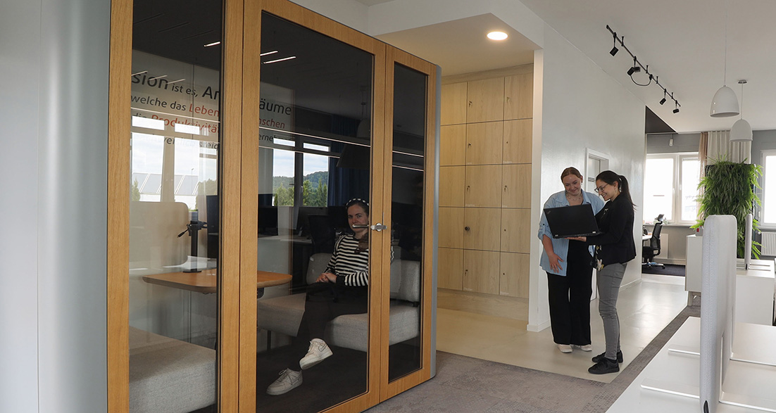 Große Akustikbox daneben zwei Personen mit Laptop im Hintergrund eine moderne Bürofläche