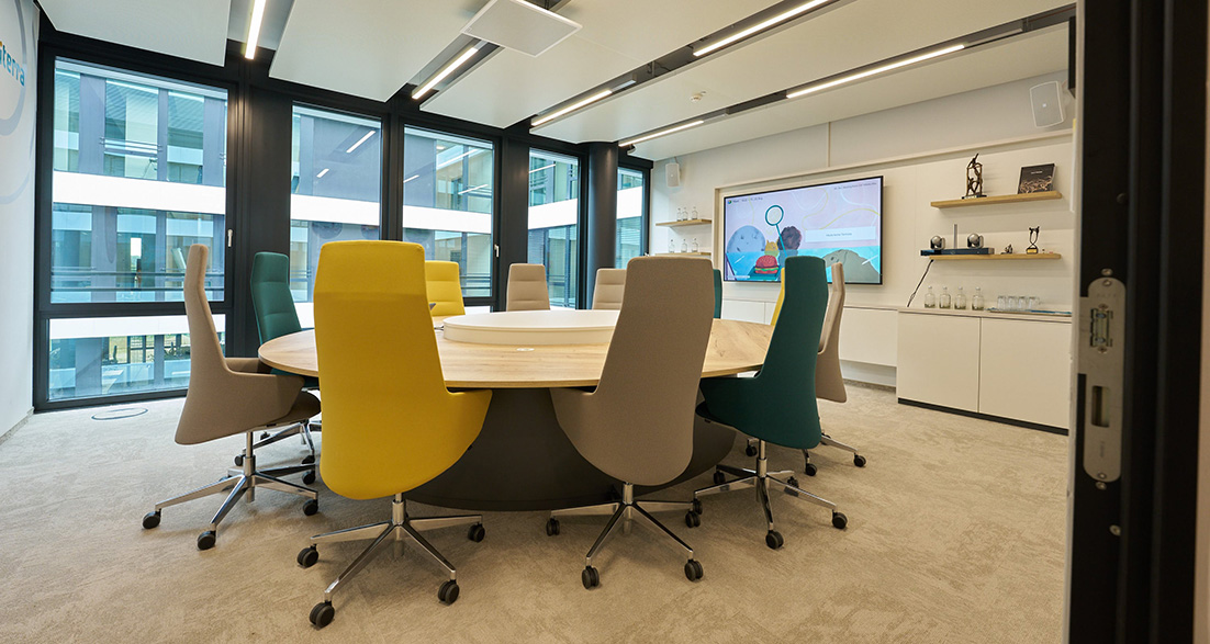Meetingraum mit rundem Tisch und bunten Konferenzstühlen und Bildschirm an Wand
