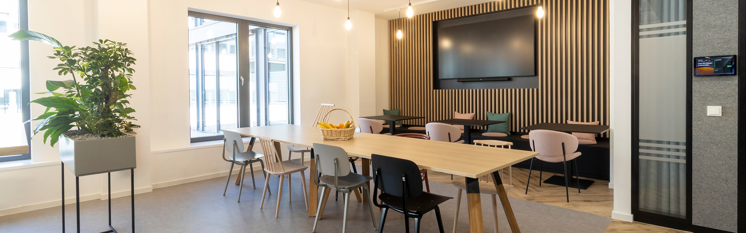 Workcafe mit Langem Tisch mit verschiedenen Stühlen und Sitzbank mit Wandbildschirm im Hintegrund