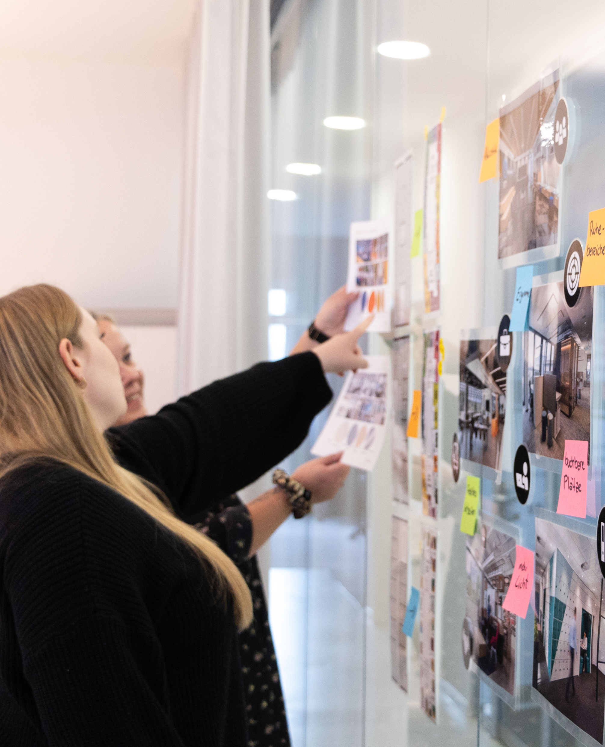 Zwei Personen zeigen auf Post Its und Bilder an einer Glaswand