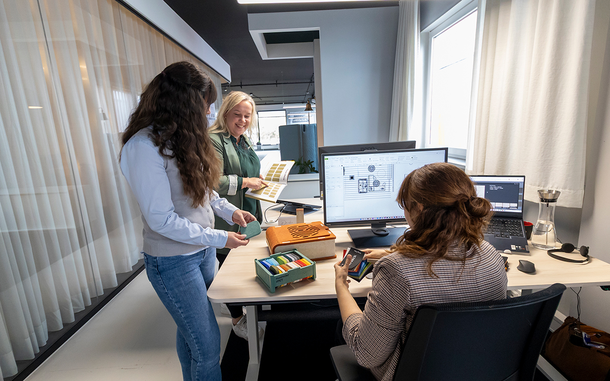 Drei Personen schauen sich Stoffmuster an zwei stehen und eine sitzt an einem Schreibtisch vor einem Bildschirm