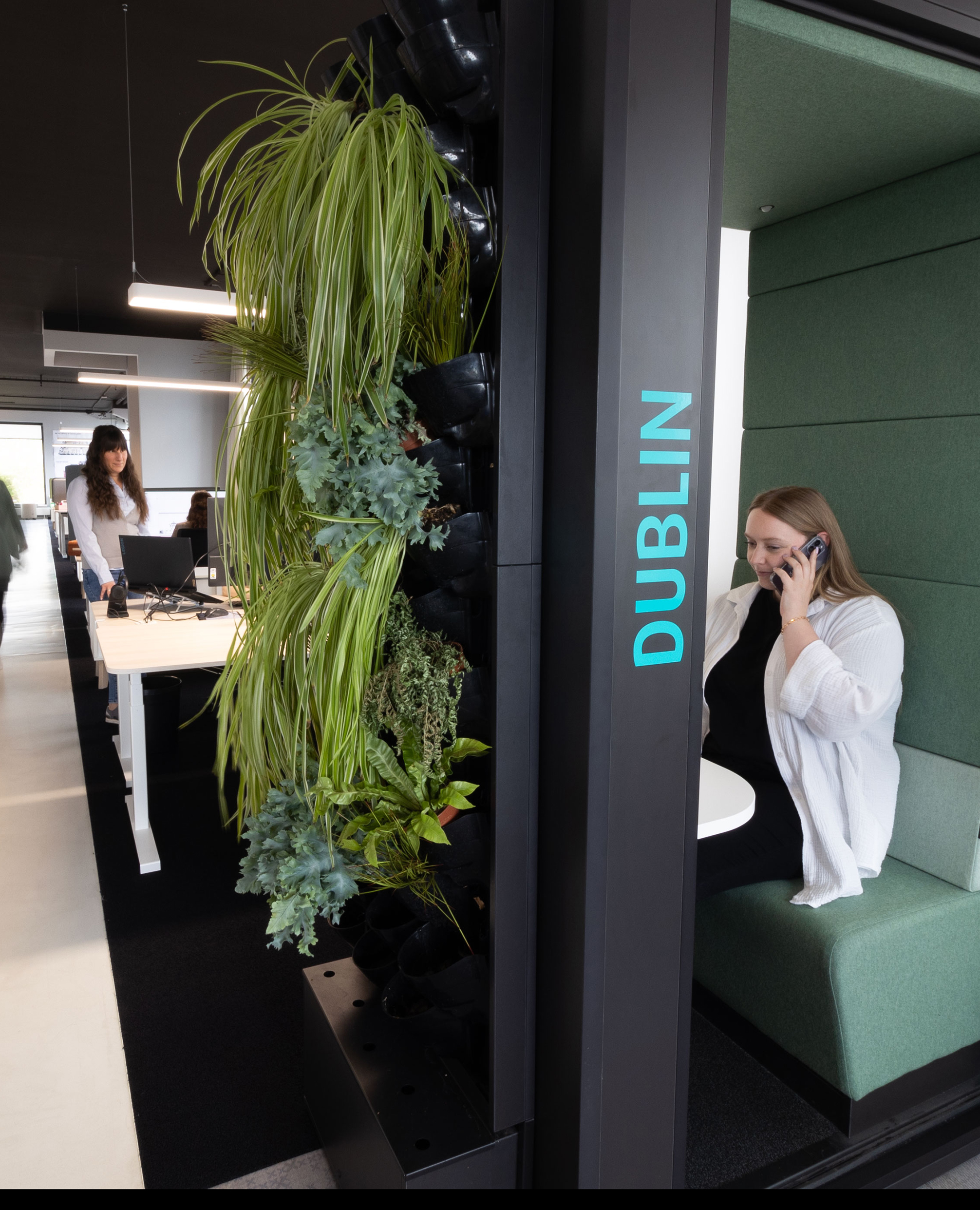 Eine Person sitzt telefonierend in einer Akustikbox im Hintergrund erkennt man weitere Personen die an einem Schreibtisch zusammen arbeiten