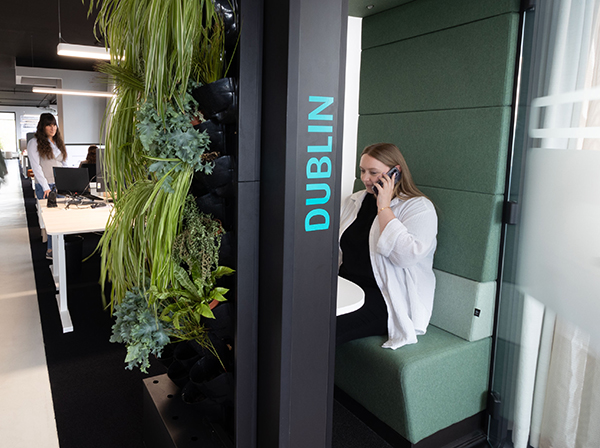 Eine Person sitzt telefonierend in einer Akustikbox im Hintergrund links arbeiten Personen am Schreibtisch