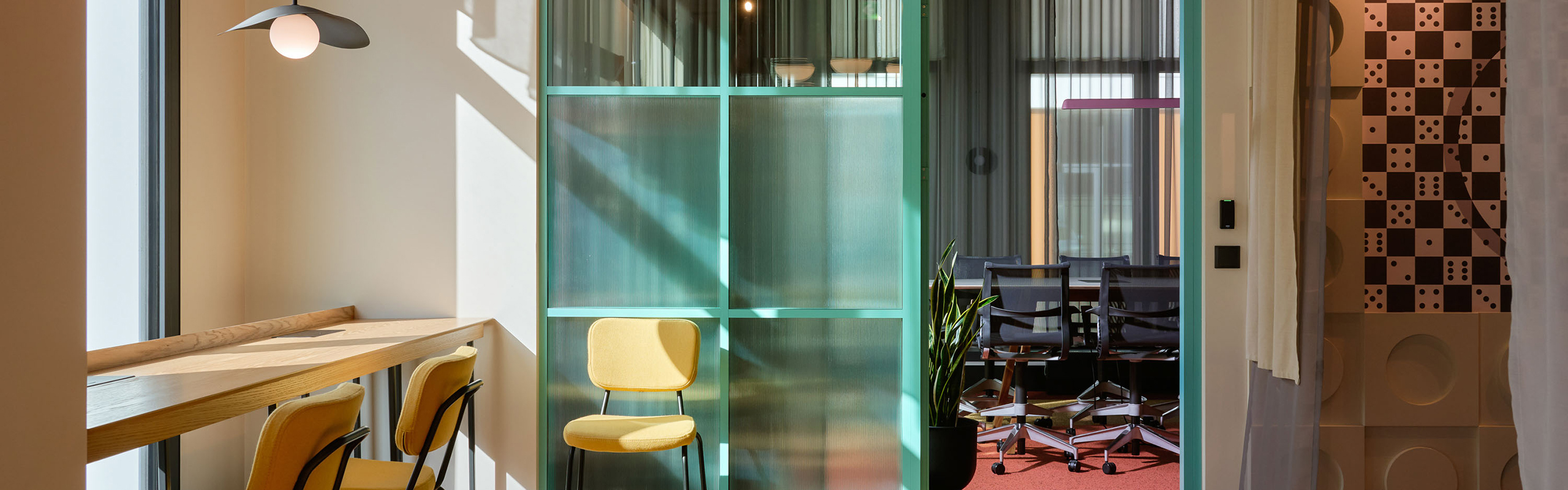 Moderner Bürobereich mit hoher Tischtheke und Barhockern am Fenster. Im Zentrum steht eine grün getönte Glaswand als Raumtrenner, dahinter ein Besprechungsraum mit mehreren Stühlen und Tisch. Warme Lichtstimmung, Pflanzen und farbliche Akzente schaffen eine offene, strukturierte Arbeitsumgebung mit unterschiedlichen Zonen.