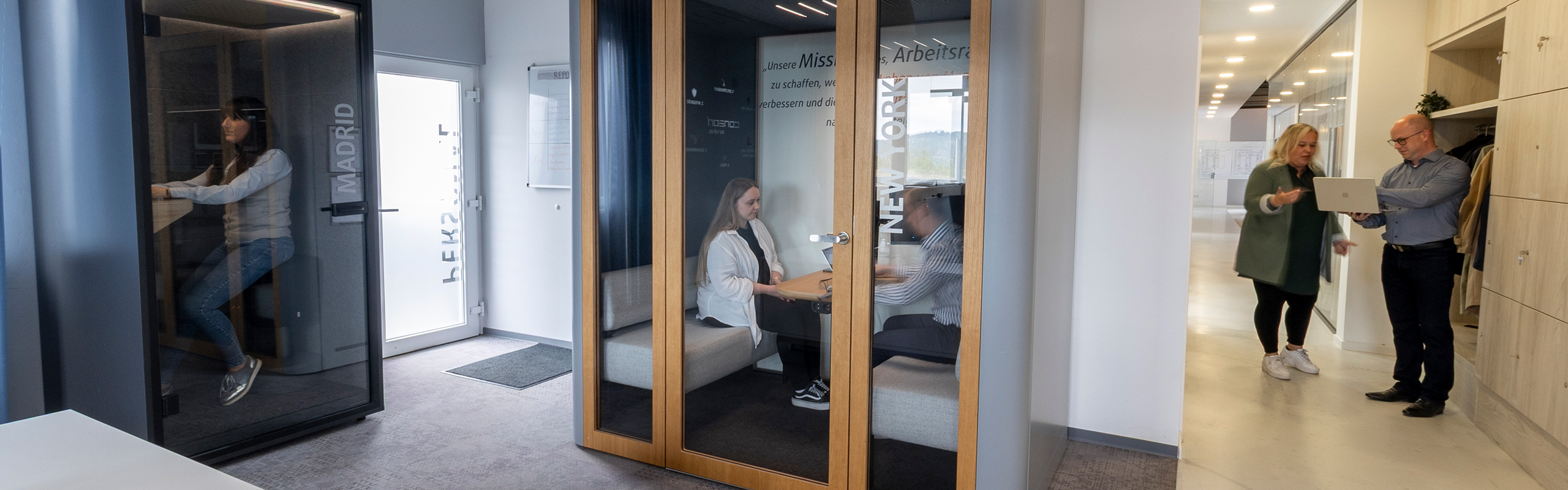 Ein modernes Büro links im Bild sitzt eine Person in einer Telefonbox in der Mitte arbeiten zwei Personen gemeinsam in einem Workpod rechts im Bild stehen zwei Personen gemeinsam an einem Laptop und diskutieren