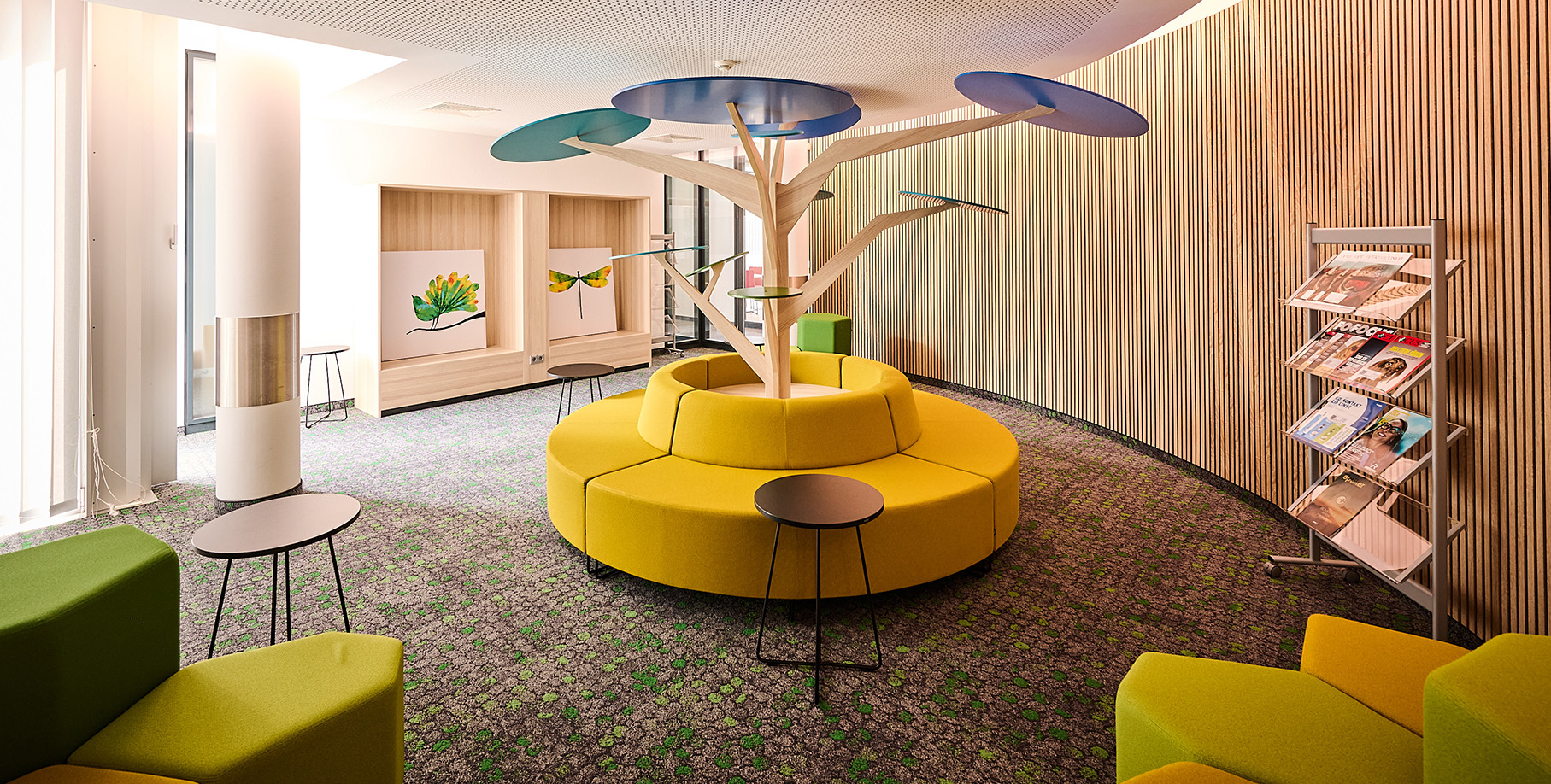 Runde Sitzbank mit Holzbaum in der Mitte und grünen und gelben Hockern im Vordergrund rechts im Bild Zeitungsauslage