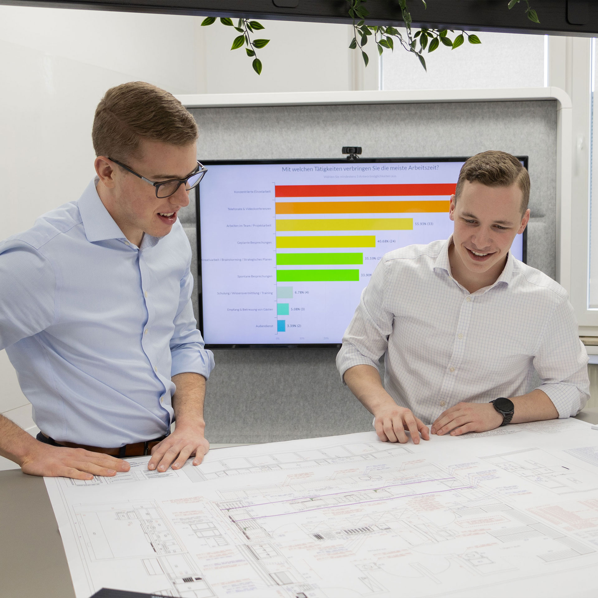 Zwei Männer mit einem Bauplan am Tisch und einer Grafik im Hintergrund