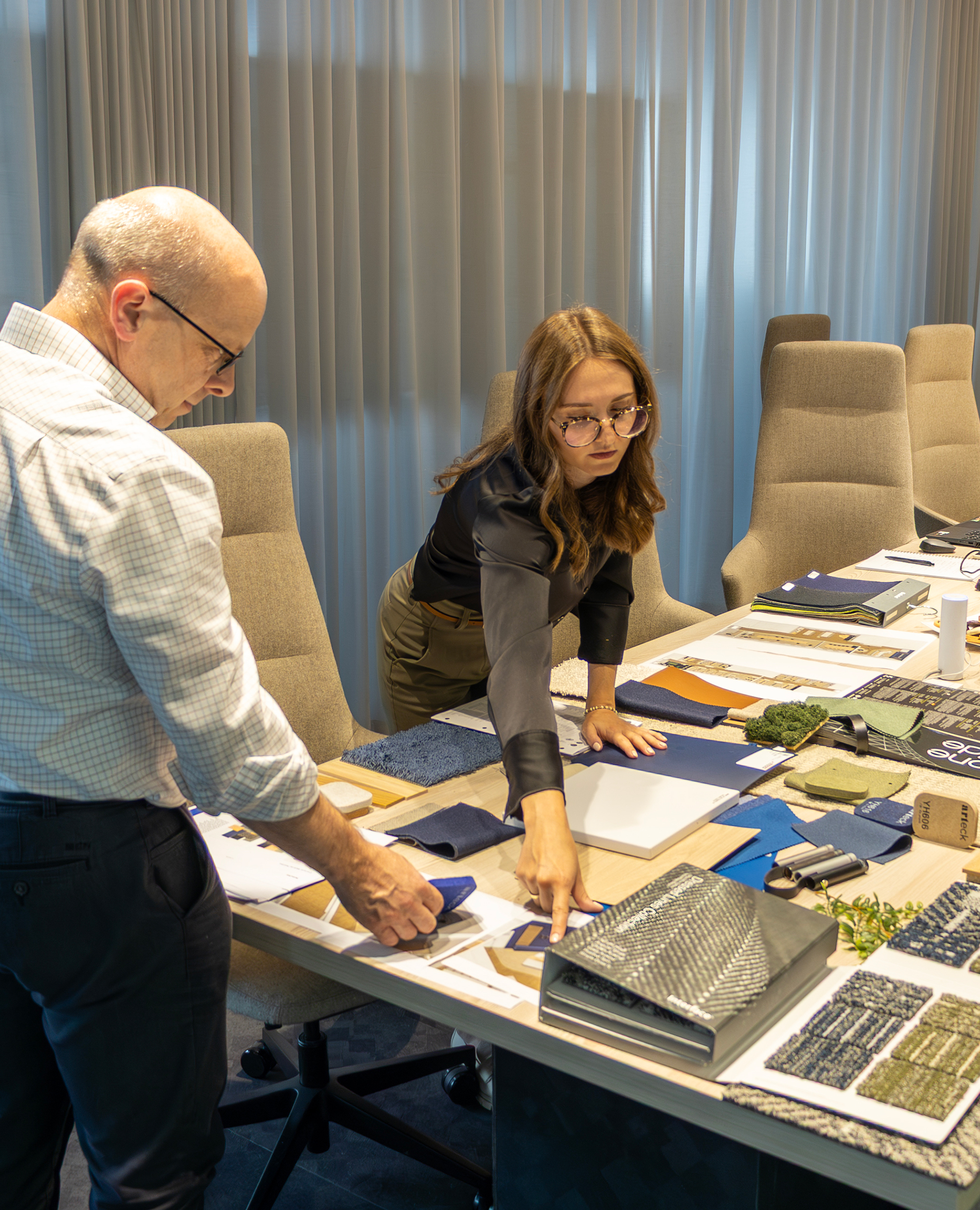 Zwei Personen stehen an einem Tisch und betrachten Material- und Farbmuster; auf dem Tisch liegen Stoffproben, Holz- und Oberflächenmuster, Musterbücher und ausgedruckte Pläne.