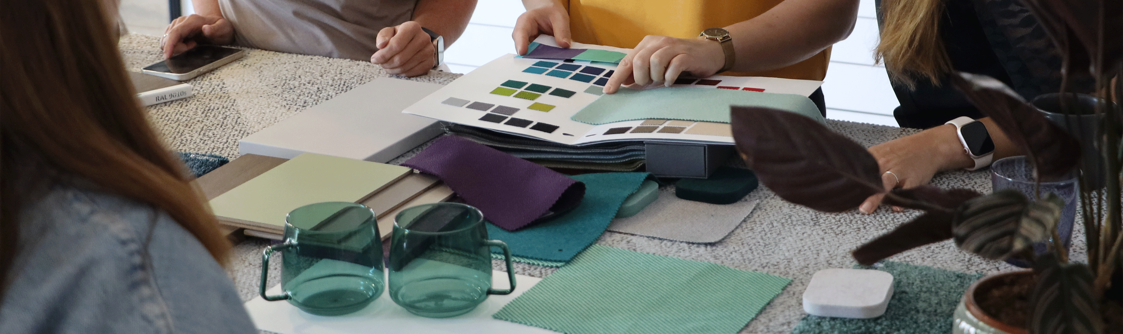 Mehrere Personen sitzen an einem Tisch und betrachten Farb- und Materialmuster; auf dem Tisch liegen Textilproben, Farbkarten, Musterbücher, Glasbecher und ein Smartphone.