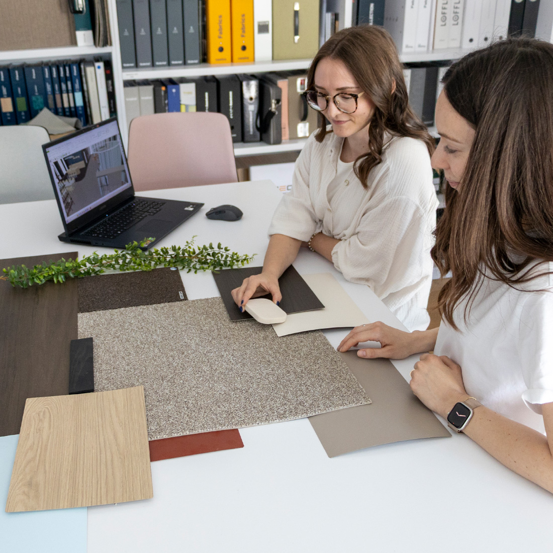Zwei Frauen sitzen an einem Tisch und betrachten Materialmuster aus Holz, Textil und Farbflächen; im Hintergrund stehen Regale mit Musterordnern und ein geöffneter Laptop.