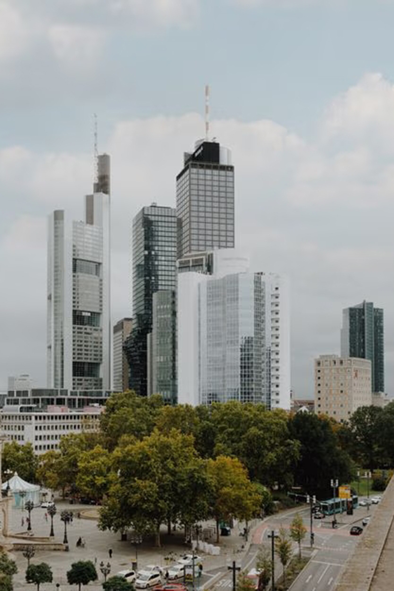 Blick auf die Frankfurter Skyline mit Hochhäusern, grünen Bäumen und Straßen im Vordergrund unter leicht bewölktem Himmel.