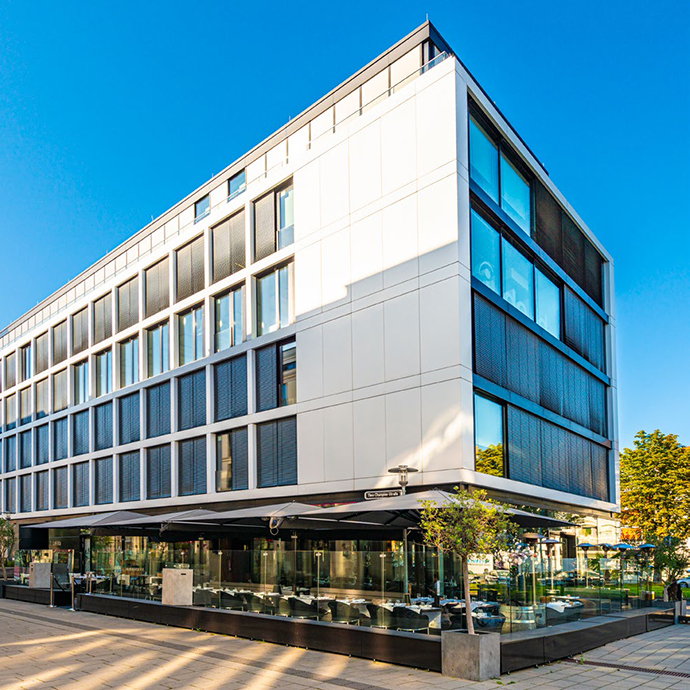 Modernes Bürogebäude mit klarer, weißer Fassadengestaltung und großen Fensterflächen an einer Straßenecke, im Erdgeschoss mit verglaster Gastronomie und Außensitzbereich, bei sonnigem Wetter.