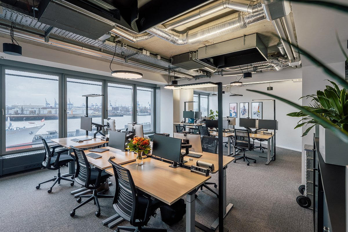 Offener Büroarbeitsbereich mit Teamarbeitsplätzen, Schreibtischen, Monitoren, sichtbarer Technikdecke und Hafenblick.