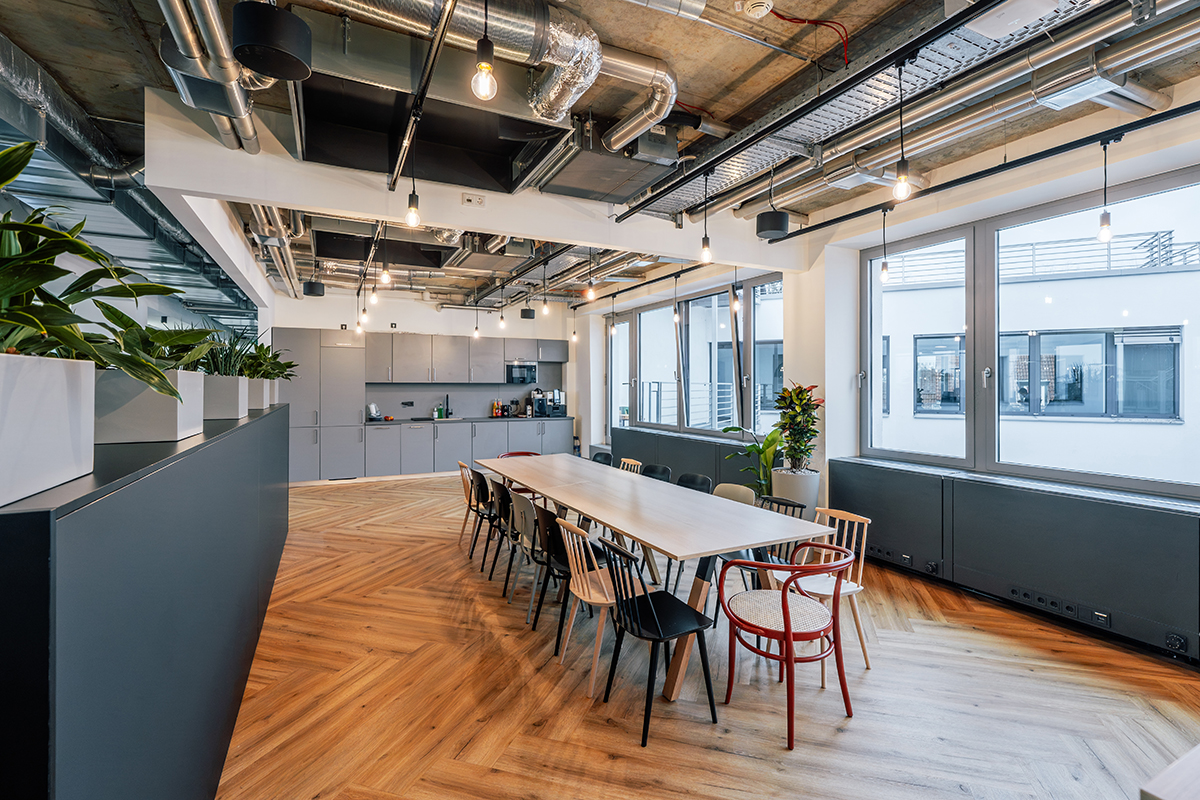 Großzügiger Aufenthalts- und Kommunikationsbereich mit langer Tischreihe, gemischten Stühlen, Pflanzen und offener Decke in moderner Bürogestaltung.