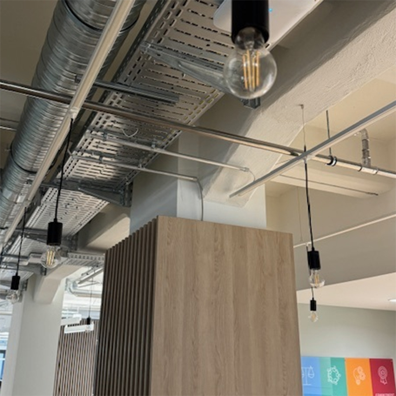 Detailaufnahme der offenen Decke mit sichtbaren Lüftungskanälen, Leuchten und Holzoberflächen.